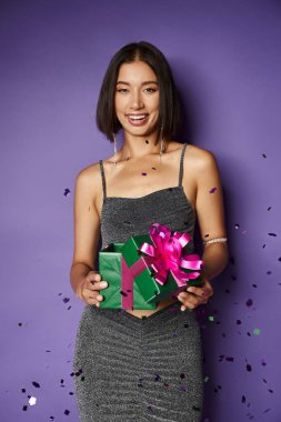 happy asian woman in party dress holding wrapped Christmas present near falling confetti on purple clipart