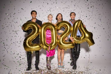 Mutlu ırklar arası arkadaşlar 2024 rakamlı balonlar tutuyorlar. Düşen konfeti yakınlarında, yeni yıl partisinde.