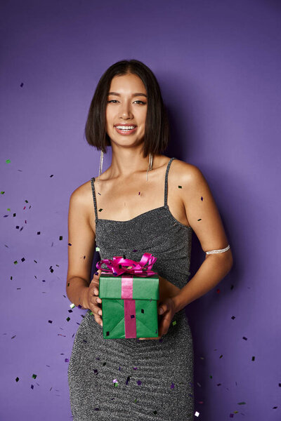 radiant asian woman in party dress holding wrapped Christmas present near falling confetti on purple