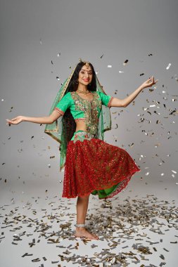 Yeşil choli ve kırmızı etekli çekici Hintli kadın konfeti yağmuru altında tek bacağının üstünde poz veriyor.