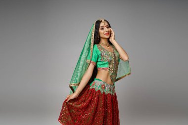 joyous indian woman in green choli touching hem of her red skirt posing with hand on cheek clipart