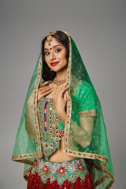 Geleneksel giysiler ve yeşil peçeyle gri zeminde poz veren Hintli genç bir kadının dikey çekimi.