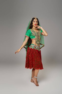 Yeşil choli ve kırmızı etekli genç, güzel Hintli kadın gri arka planda poz veriyor.
