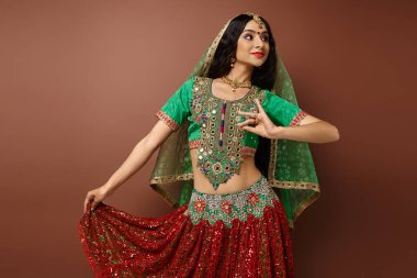 Geleneksel giyinmiş Bindi 'li çekici Hintli kadın kahverengi arka planda dans ederken el kol hareketi yapıyor.