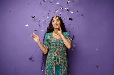 beautiful amazed indian woman posing under confetti rain on purple backdrop with hand near face clipart