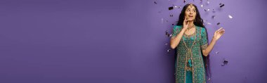 surprised young indian woman under confetti rain on purple backdrop with hand near face, banner clipart