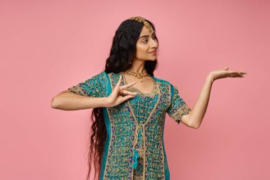 Ulusal kostüm giymiş genç ve çekici Hintli kadın pembe arka planda el kol hareketi yapıyor ve başka tarafa bakıyor.