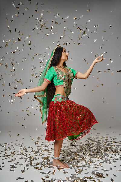 vertical shot of pretty indian woman in national attire posing on one leg under confetti rain