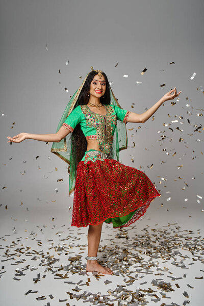 attractive young indian woman in green choli and red skirt posing on one leg under confetti rain