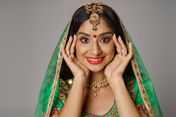 cheerful young indian woman with bindi dot and green veil smiling at camera with hands near face