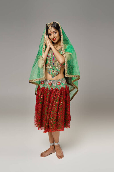 joyful indian woman in red skirt and green choli posing on gray backdrop and looking at camera