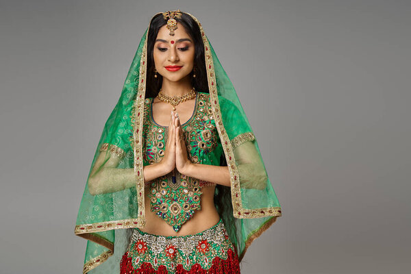 joyous young indian woman in traditional costume with veil and bindi praying and looking down
