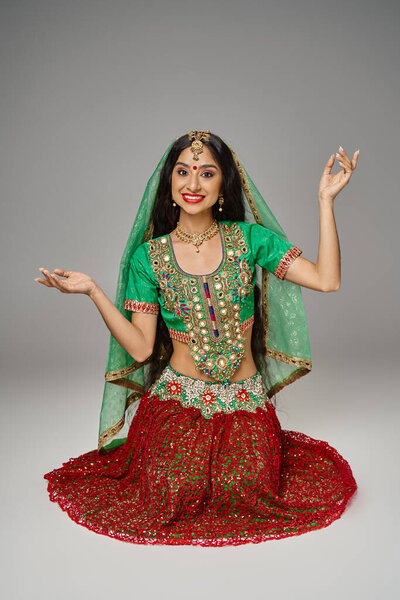 joyous young indian woman in red skirt and green choli gesturing and smiling happily at camera