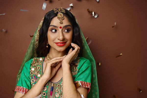cheerful indian woman in traditional attire with bindi dot on forehead standing under confetti rain