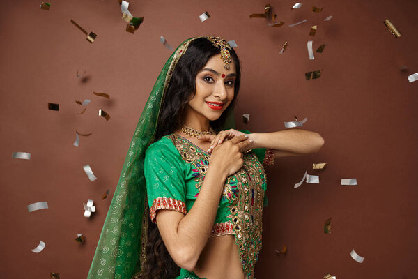 beautiful indian woman in green choli with red skirt with veil and bindi posing under confetti rain