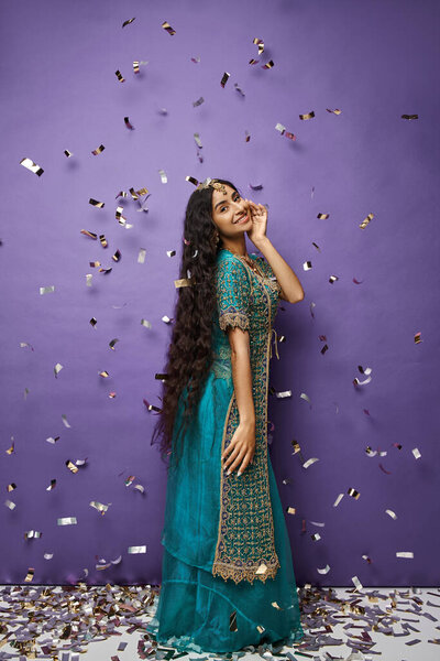 привлекательная веселая индийская женщина в синем сари позирует под дождём конфетти на фиолетовом фоне