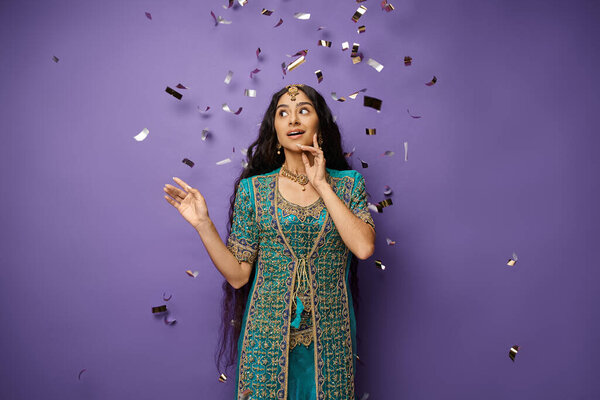 красивая изумленная индианка позирует под дождём конфетти на фиолетовом фоне с рукой рядом с лицом