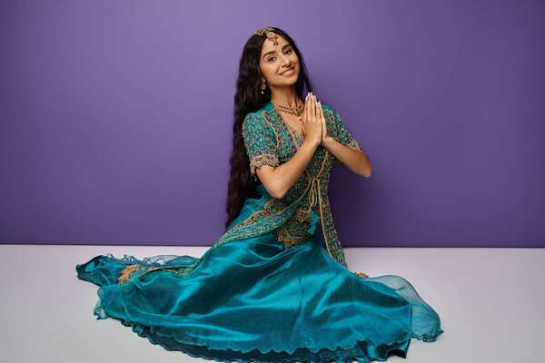 Радостная индийская женщина сидит на полу на фиолетовом фоне и молится, улыбаясь в камеру.