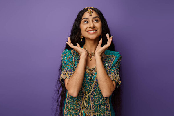 happy indian woman with long hair in blue sari posing on purple backdrop with hands near face