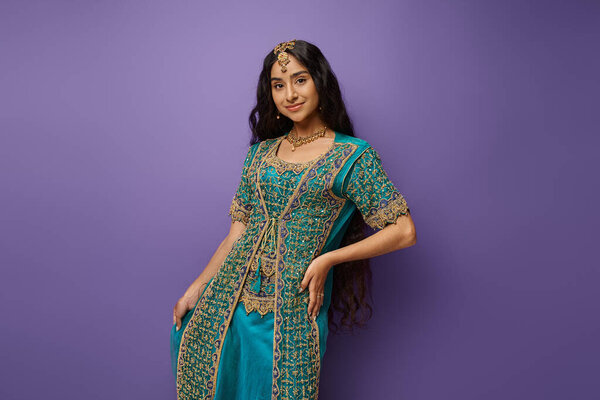 good looking young indian woman in traditional blue sari posing with hand on hip on purple backdrop