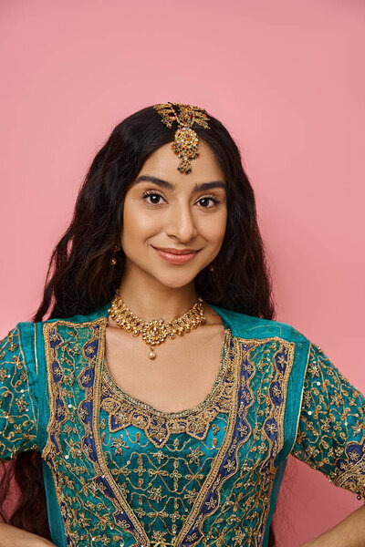 vertical shot of young indian woman in blue sari with accessories smiling at camera on pink backdrop
