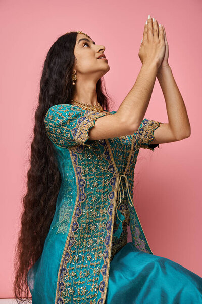 vertical shot of indian woman in blue sari showing praying gesture looking away and on pink backdrop