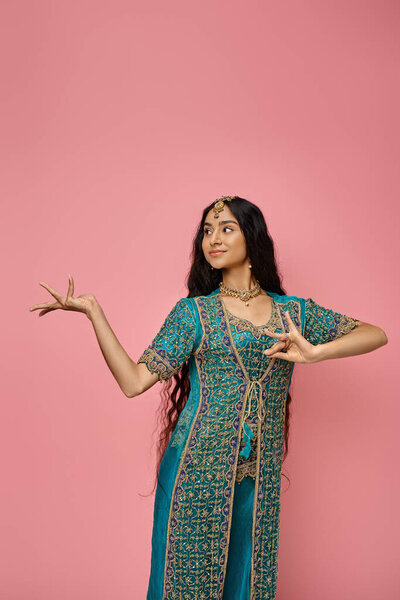 joyful attractive indian woman in traditional blue sari posing on pink background and looking away