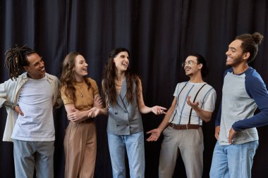 cheerful multicultural colleagues posing and laughing near black drape in office, group photo clipart