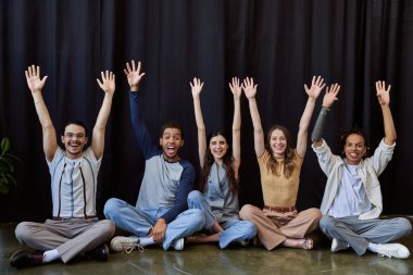 excited stylish multiethnic team sitting and waving hands near black drape in office, group photo clipart