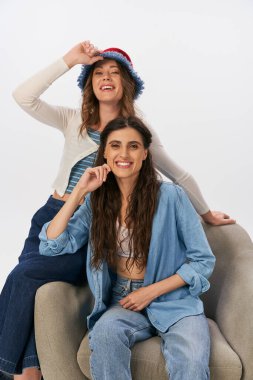 Kot elbiseli neşeli kadın, gri arka planda panama şapkalı kız arkadaşının yanında koltukta oturuyor.