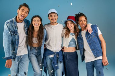 Moda kıyafetleri içinde neşeli ırklar arası arkadaşlar konfeti yağmurunun altında mavi renkli kameraya bakıyorlar.