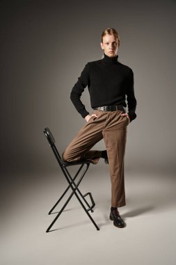 attractive non binary model with ponytail posing with knee on black chair and looking at camera clipart