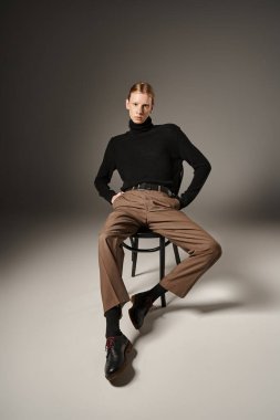 Siyah koltukta elleri ceplerinde oturan çekici, genç, ikilik olmayan kişinin dikey görüntüsü.