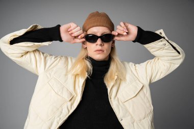 Güneş gözlüklerine dokunan ve kameraya bakan şapkalı, şık görünümlü, ikili olmayan model.