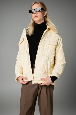 Gri arka planda duran güneş gözlüklü, modaya uygun, sıcak giyimli, genç ve çekici olmayan model.