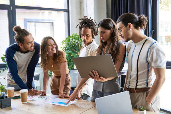 female team lead with laptop pointing at graphs on meeting with creative interracial team in office