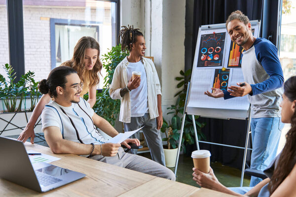 веселый африканский американец, говорящий рядом с флип-чартом во время встречи с многонациональной командой в офисе