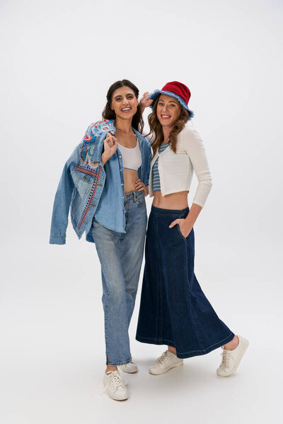счастливая женщина в панамской шляпе позирует с рукой в кармане рядом с девушкой в джинсовой одежде на сером фоне