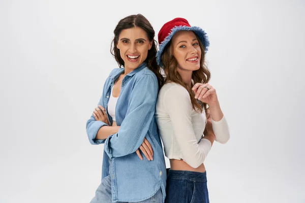 stock image happy women in jeans wear and panama hat posing back to back with folded arms on grey, fashion