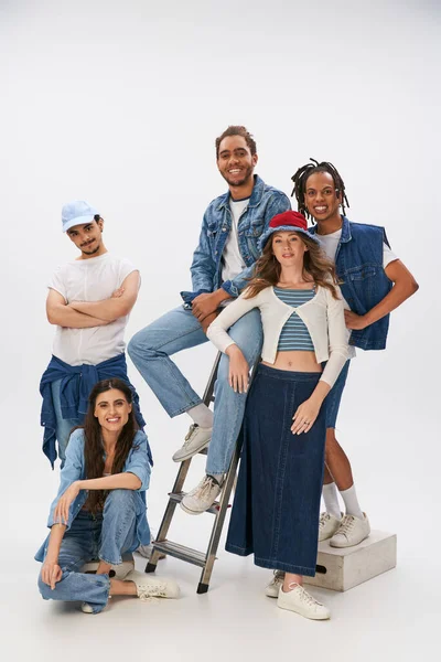 happy multiethnic friends in trendy street wear posing with ladder and smiling at camera on grey