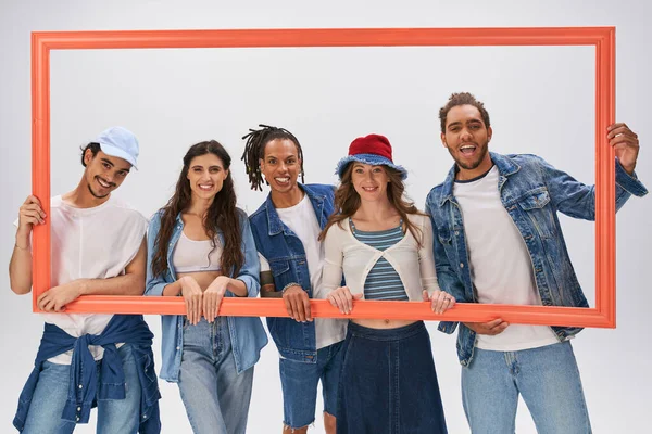 group portrait of stylish interracial friends posing with picture frame on grey backdrop, fashion