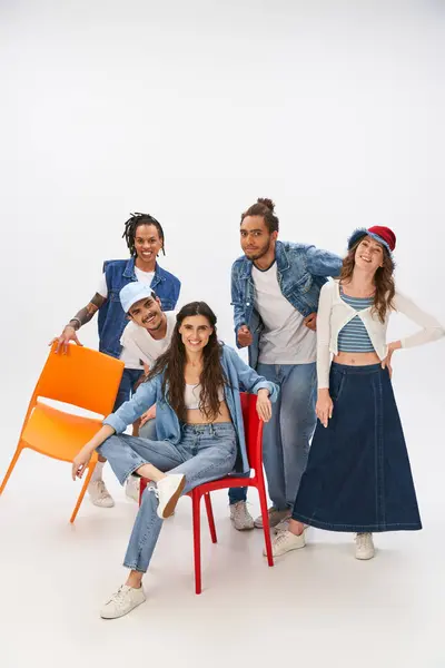 smiling woman sitting on chair near multiethnic friends in trendy casual attire on grey backdrop