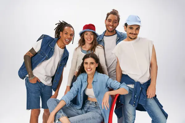 stock image woman in denim wear sitting on red chair and smiling at camera near  interracial friends on grey