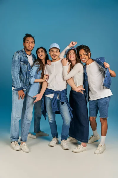 group portrait of interracial friends in trendy and denim clothes on blue backdrop, full length