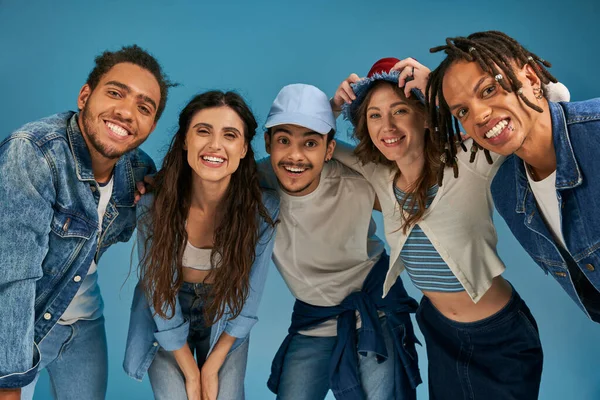 cheerful multiethnic friends in trendy street wear smiling at camera om blue backdrop, urban style