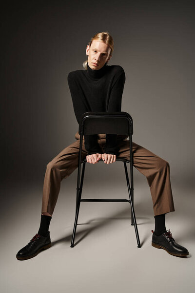 attractive non binary person in b lack turtleneck and brown pants sitting on black chair, fashion