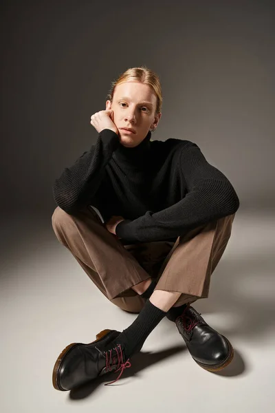 good looking young non binary model sitting on floor with crossed legs and hand near face, fashion