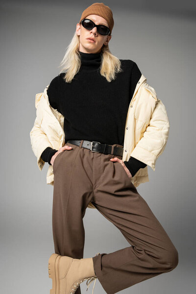 красивая не бинарная модель в шляпе шапочки и солнцезащитных очках, позируя руками в карманах, мода