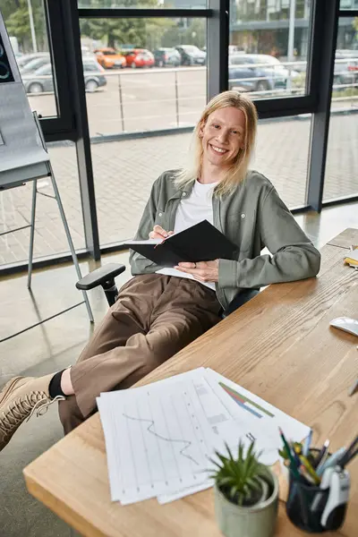 cheerful non binary person sitting at desk with notebook in hands and smiling at camera, business