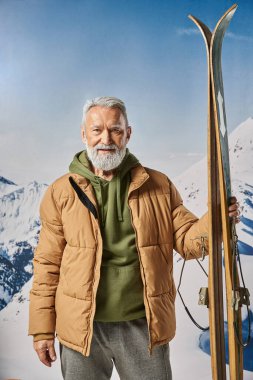 athletic man dressed as Santa holding ski looking at camera with mountains backdrop, winter concept clipart